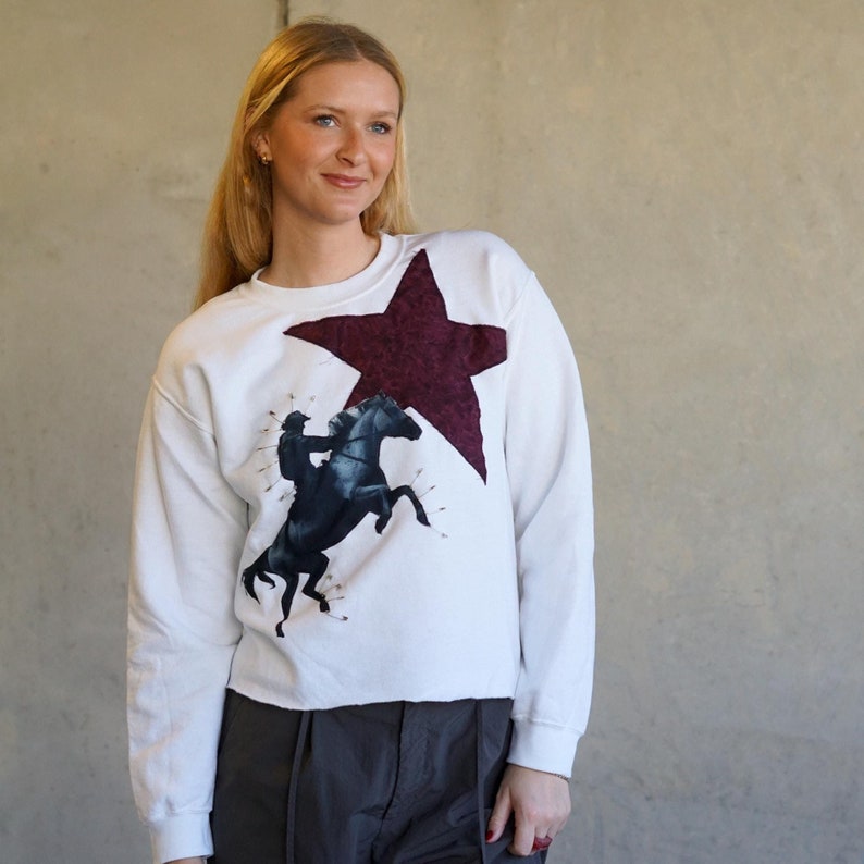 May include: White sweatshirt featuring a large burgundy star and a graphic of a rider on a rearing black horse. The sweatshirt has a crew neck and long sleeves. The design is centered on the front of the shirt.