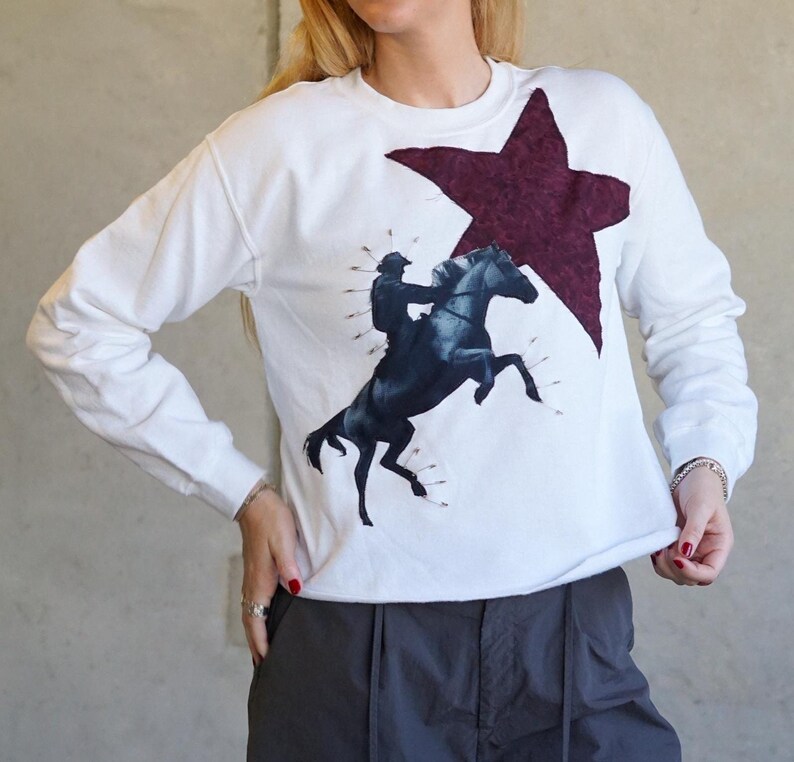 May include: White long-sleeve sweatshirt featuring a dark blue graphic of a rider on a rearing horse, pierced with arrows. A large burgundy star is above the horse. The sweatshirt has a crew neck and is made of a soft material.