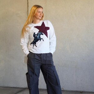 May include: White sweatshirt featuring a maroon star and a graphic of a rider on a rearing black horse. The sweatshirt is paired with dark gray cargo pants. The outfit is casual and suitable for everyday wear.