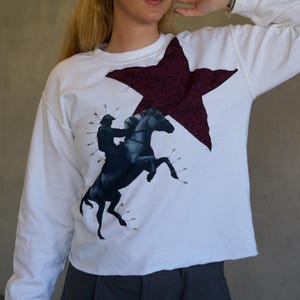 May include: White long-sleeve sweatshirt featuring a large burgundy star and a black and white graphic of a rider on a rearing horse. The sweatshirt has a raw hemline and a relaxed fit, suitable for casual wear.