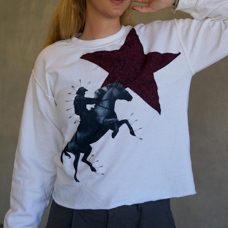 May include: White long-sleeve sweatshirt featuring a large burgundy star and a black and white graphic of a rider on a rearing horse. The sweatshirt has a raw hemline and a relaxed fit, suitable for casual wear.