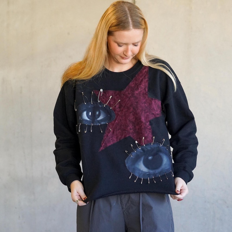 May include: Black sweatshirt with a large red star and two eyes appliqued on the front. The eyes are surrounded by safety pins.