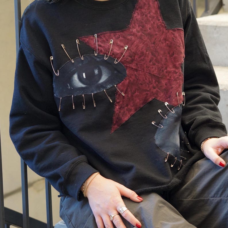 May include: Black sweatshirt featuring a red star and an eye design, both secured with safety pins.