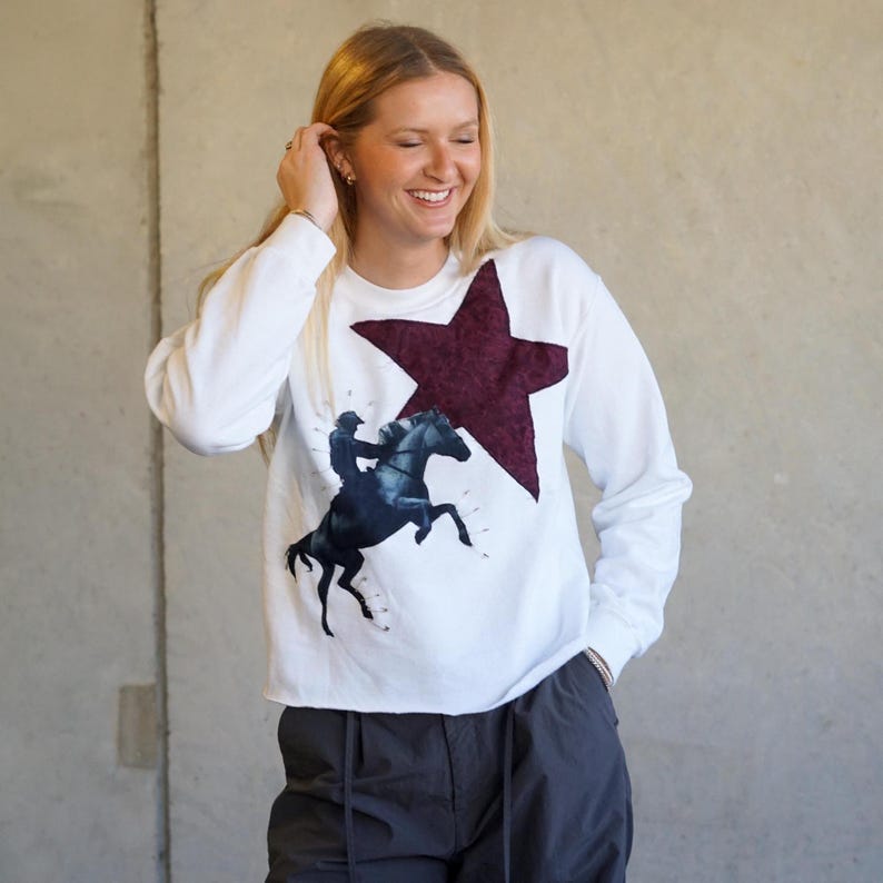 May include: White long-sleeve sweatshirt featuring a large burgundy star and a graphic of a rider on a rearing horse. The sweatshirt has a crew neck and a relaxed fit, suitable for casual wear.