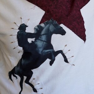 May include: White fabric with a black and gray image of a rider on a rearing horse, stitched onto the fabric. A burgundy star is in the background. Safety pins are used to attach the image to the fabric.