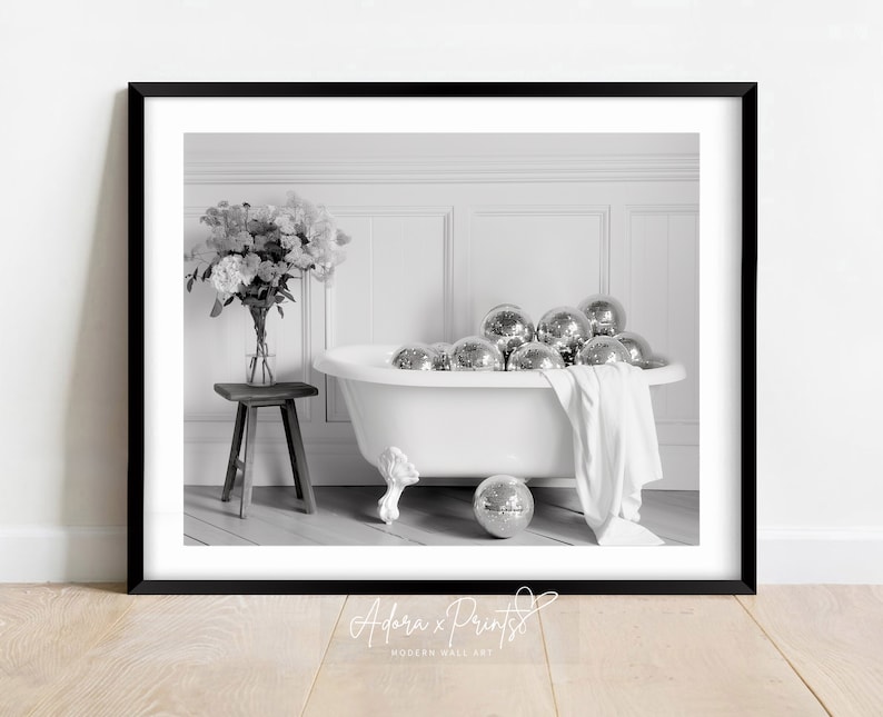 Pode incluir: Uma fotografia a preto e branco de uma banheira branca cheia de bolas de discoteca prateadas. Um ramo de flores est&aacute; sobre um pequeno banquinho de madeira ao lado da banheira. A imagem est&aacute; emoldurada por uma moldura preta.