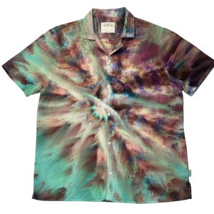 May include: Short-sleeved button-down shirt with a unique tie-dye pattern in shades of teal, brown, and purple. The shirt features a camp collar and a label near the collar.