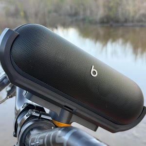 May include: A black Beats Pill+ portable Bluetooth speaker mounted on a bicycle handlebar. The speaker has a black mesh grille and a silver Beats logo.