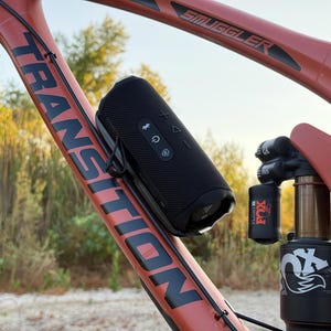 May include: A black portable speaker is mounted on a red mountain bike frame. The bike frame has the word "TRANSITION" printed on it. The speaker has control buttons and is attached with a black strap. The bike's suspension system is also visible.