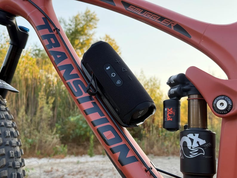May include: A black portable speaker is mounted on a red mountain bike frame. The bike frame has the word "TRANSITION" printed on it. The speaker has control buttons and is attached with a black strap. The bike's suspension system is also visible.