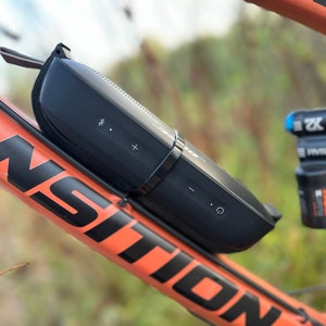 Puede incluir: Un altavoz Bluetooth portátil negro fijado a un cuadro de bicicleta de montaña naranja brillante. El altavoz tiene botones de control de volumen y un botón de encendido.