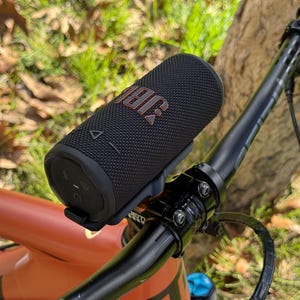 May include: A black JBL portable speaker mounted on a bicycle handlebar. The speaker has the JBL logo in orange. The bicycle is orange and black, with a black handlebar and a green background.