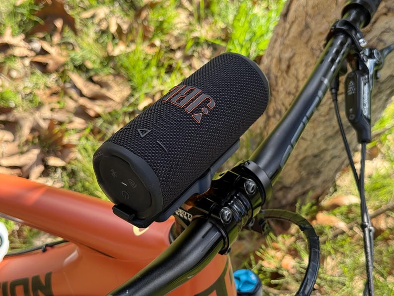 May include: A black JBL portable speaker mounted on a bicycle handlebar. The speaker has the JBL logo in orange. The bicycle is orange and black, with a black handlebar and a green background.