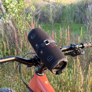 May include: A black cylindrical portable speaker is mounted on the handlebars of a mountain bike. The speaker is secured with a black strap. The bike has an orange frame and black handlebars with textured grips. The background features green and brown foliage.