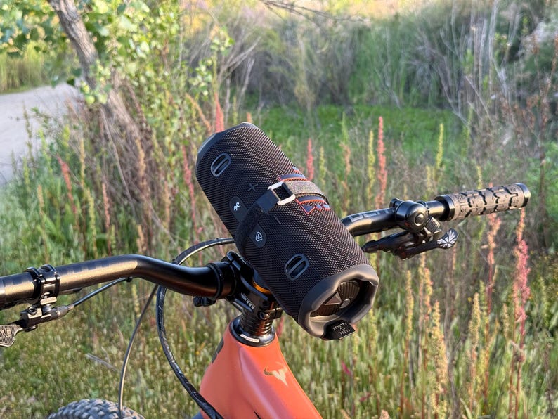 May include: A black cylindrical portable speaker is mounted on the handlebars of a mountain bike. The speaker is secured with a black strap. The bike has an orange frame and black handlebars with textured grips. The background features green and brown foliage.
