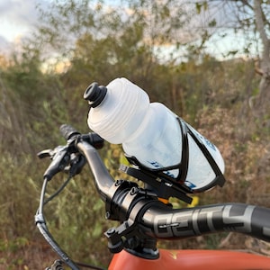 May include: A black bicycle handlebar with a clear water bottle attached to a black cage. The bottle is partially filled with water. The handlebar has a black grip with the word "DEITY" printed on it.