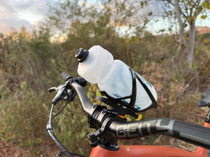 May include: A black bicycle handlebar with a clear water bottle attached to a black cage. The bottle is partially filled with water. The handlebar has a black grip with the word "DEITY" printed on it.