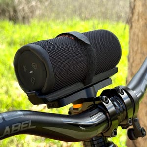 May include: A black cylindrical speaker mounted on a bicycle handlebar. The speaker is secured with a black strap and sits on a black bracket. The handlebar is black with the word "BLACKLABEL" printed on it. The background is blurred green.