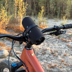 May include: A black portable speaker is mounted on the handlebars of an orange mountain bike. The bike has black handlebars and a black seat. The speaker is secured with a black strap. The background shows dry grass and a dirt path.