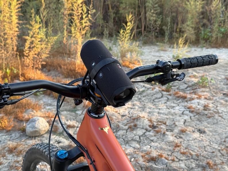 May include: A black portable speaker is mounted on the handlebars of an orange mountain bike. The bike has black handlebars and a black seat. The speaker is secured with a black strap. The background shows dry grass and a dirt path.