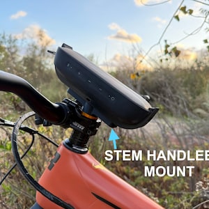 May include: A black speaker mounted on a bicycle handlebar stem. The speaker is attached to the stem with a black mount. The bicycle is orange and has a black handlebar.