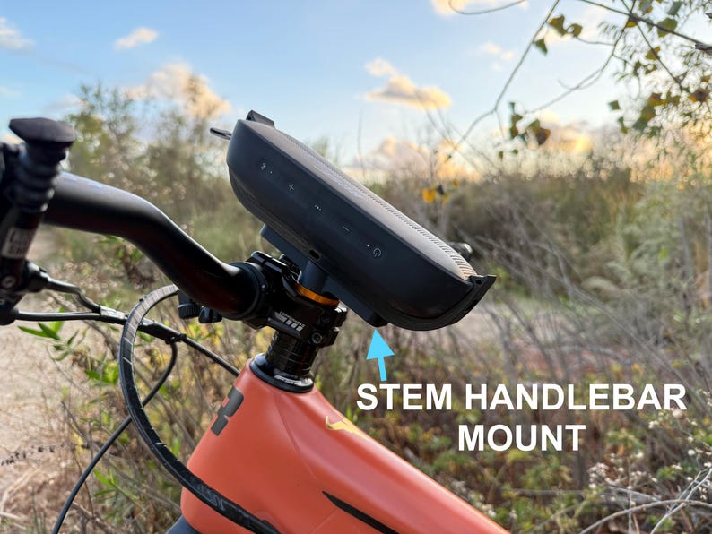 May include: A black speaker mounted on a bicycle handlebar stem. The speaker is attached to the stem with a black mount. The bicycle is orange and has a black handlebar.