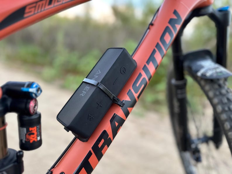 May include: A black portable Bluetooth speaker attached to the frame of a red mountain bike. The speaker has volume controls and a power button.