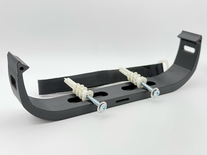 May include: Black plastic wall mount bracket with two white plastic anchors and screws. The bracket has a curved shape and is designed to hold a pipe or cable.