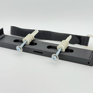 May include: Black plastic wall mount with two white plastic anchors and screws. The mount has a black strap with a buckle.