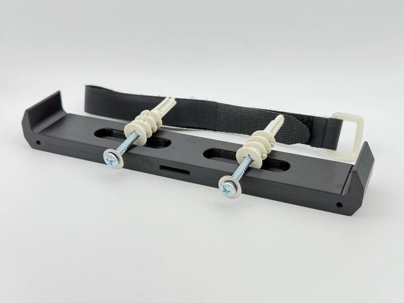 May include: Black plastic wall mount with two white plastic anchors and screws. The mount has a black strap with a buckle.