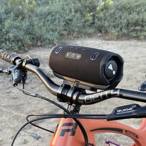 May include: A black cylindrical portable speaker mounted on the handlebars of an orange mountain bike. The speaker has control buttons and a triangular logo. The bike's handlebars are black, and the frame is orange.