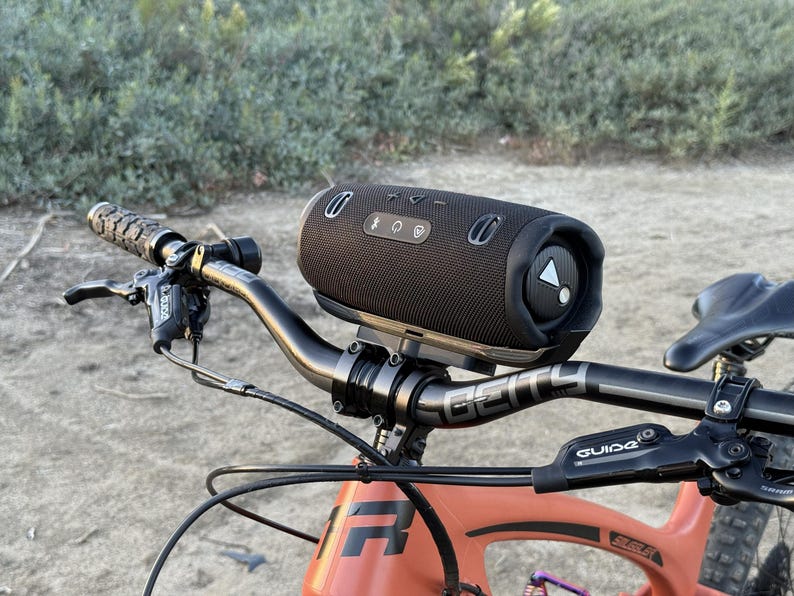 May include: A black cylindrical portable speaker mounted on the handlebars of an orange mountain bike. The speaker has control buttons and a triangular logo. The bike's handlebars are black, and the frame is orange.