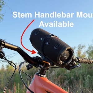 May include: A black cylindrical speaker is mounted on the handlebars of an orange mountain bike. The text "Stem Handlebar Mount Available" is displayed above the speaker with a red arrow pointing to the mount. The bike is set against a backdrop of a blue sky and green foliage.
