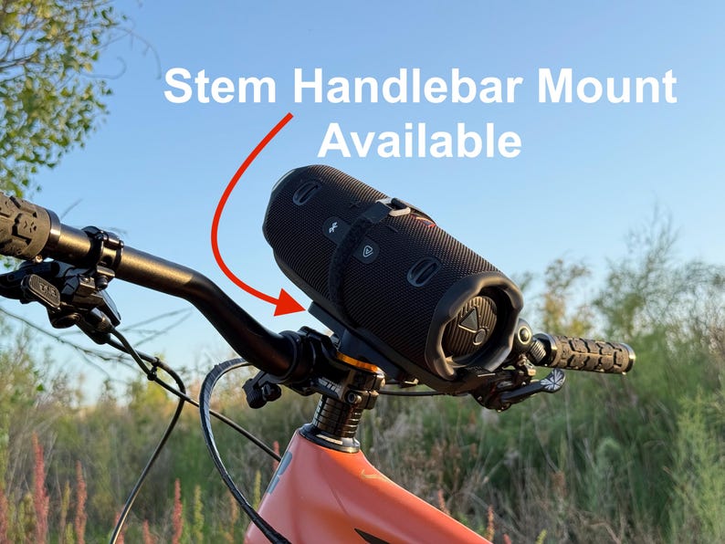 May include: A black cylindrical speaker is mounted on the handlebars of an orange mountain bike. The text "Stem Handlebar Mount Available" is displayed above the speaker with a red arrow pointing to the mount. The bike is set against a backdrop of a blue sky and green foliage.