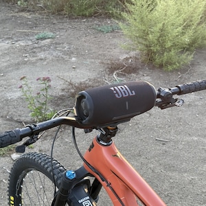 May include: A black JBL speaker mounted on the handlebars of an orange mountain bike. The speaker is cylindrical with the JBL logo in white. The bike has black handlebars and a black tire.