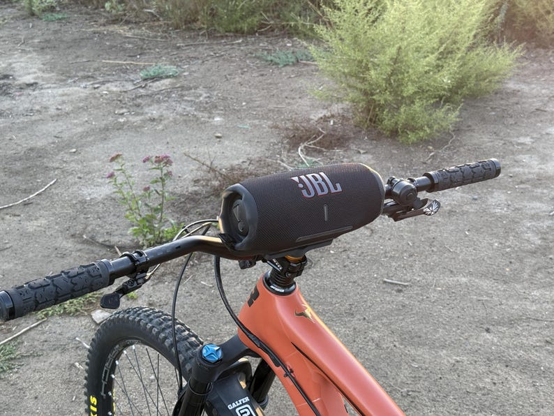 May include: A black JBL speaker mounted on the handlebars of an orange mountain bike. The speaker is cylindrical with the JBL logo in white. The bike has black handlebars and a black tire.