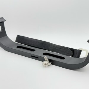 May include: A black plastic bracket with a black strap and a silver screw. The bracket has three slots and is designed to hold an object securely.