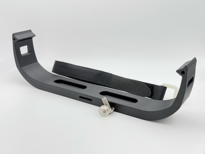 May include: A black plastic bracket with a black strap and a silver screw. The bracket has three slots and is designed to hold an object securely.