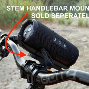 May include: A black portable speaker mounted on a bicycle handlebar. The speaker is cylindrical with control buttons and a mesh cover. The text "STEM HANDLEBAR MOUNT SOLD SEPERATELY" is visible above the speaker.