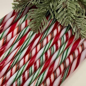 May include: A collection of long, cylindrical candy canes arranged in a parallel row, each featuring a repeating diagonal pattern of red, green, and white stripes. A sprig of green pine needles rests across the top of the candy canes.