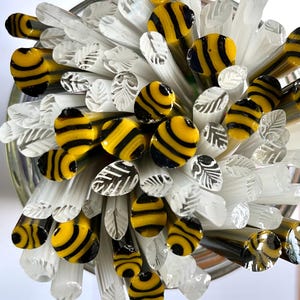 May include: Close-up of a collection of glass straws. Some straws are clear with leaf-like designs, while others feature a yellow and black striped pattern resembling bees. The straws are arranged in a circular shape.