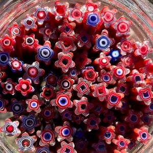 May include: A close-up of a jar filled with red, white, and blue star-shaped glass beads. The beads are made of layered glass, creating a swirling pattern.