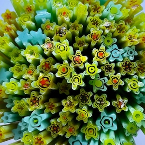 May include: Close-up of a collection of colorful glass rods. The rods are primarily yellow and green, with star-shaped and floral designs in red, black, and white. Some rods are blue. The ends of the rods are visible, creating a textured, patterned surface.