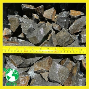 May include: A collection of dark grey and brown jagged rocks piled together. A yellow measuring tape is placed across the stones, showing a span of approximately 10 inches, with markings ranging from 1 to 11 inches and 4 to 29 centimeters.