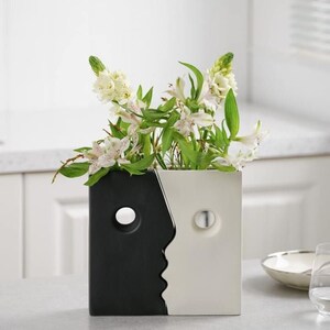 May include: A black and white ceramic vase with a face design. The vase has two cut-out circles for eyes. The vase is filled with white flowers.