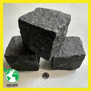 May include: Three dark gray granite blocks with rough, textured surfaces. The blocks are cube-shaped and vary slightly in size. A coin is next to the blocks. The Earth Depot logo is in the lower left corner.