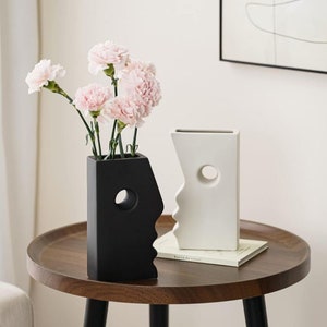 Puede incluir: Dos jarrones de cer&aacute;mica con forma de cara, uno negro y otro blanco, con un agujero en forma de ojo. Los jarrones est&aacute;n sobre una mesa de madera con un libro y claveles rosas en el jarr&oacute;n negro.