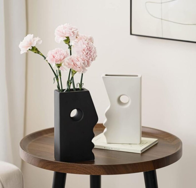 Puede incluir: Dos jarrones de cer&aacute;mica con forma de cara, uno negro y otro blanco, con un agujero en forma de ojo. Los jarrones est&aacute;n sobre una mesa de madera con un libro y claveles rosas en el jarr&oacute;n negro.