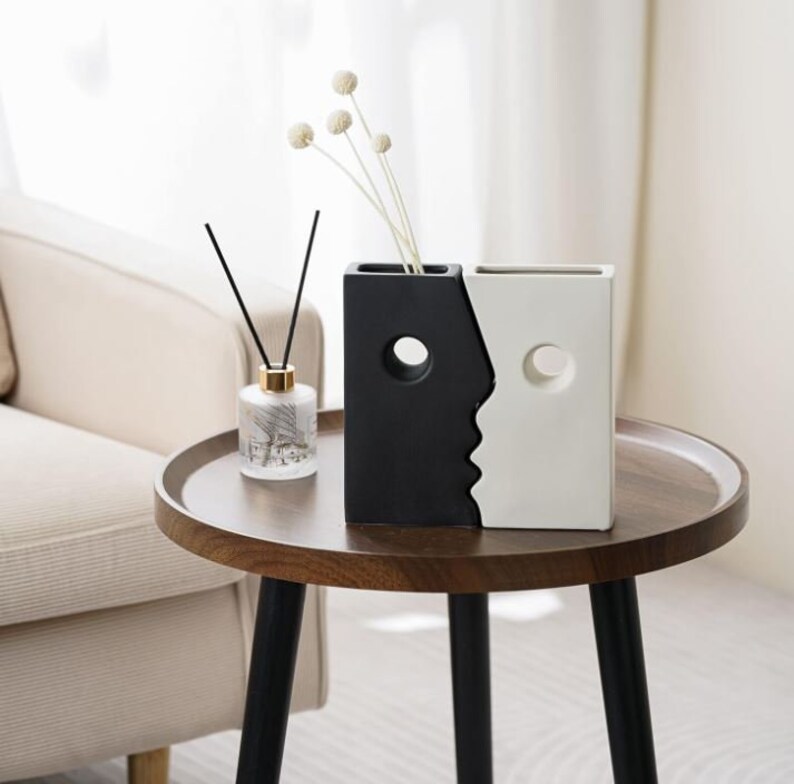 Puede incluir: Un jarr&oacute;n de cer&aacute;mica blanco y negro con un dise&ntilde;o de cara. El jarr&oacute;n tiene dos aberturas para flores. El jarr&oacute;n est&aacute; sobre una mesa de madera.
