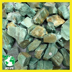 May include: A pile of rough-cut, green and tan Amazonite stones. The stones vary in size and shape, with visible textures and natural patterns. A small Earth Depot logo is in the lower left corner.